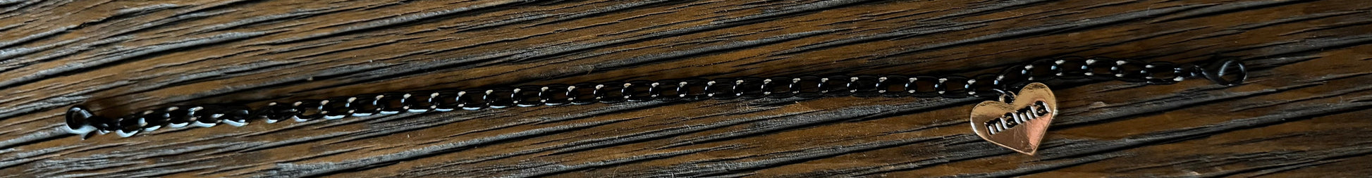 A black metal chain bracelet with a small heart-shaped charm engraved with the MAMA, displayed on a dark wood background.