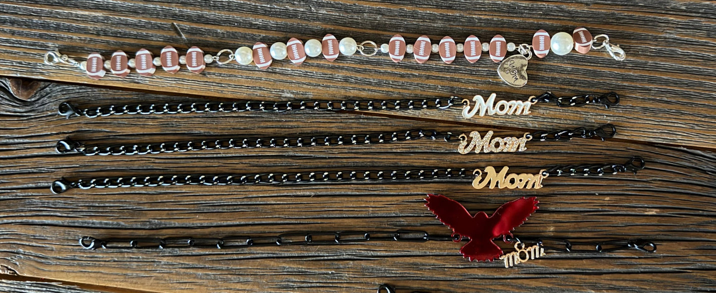 A collection of personalized mother's day bracelets laid out on a rustic wooden plank background. The bracelets include black beaded chains, black links with gold 'Mom' charms, a red velvet ribbon with gold 'Mom' charms, and a pink and white beaded bracelet with a small heart charm.