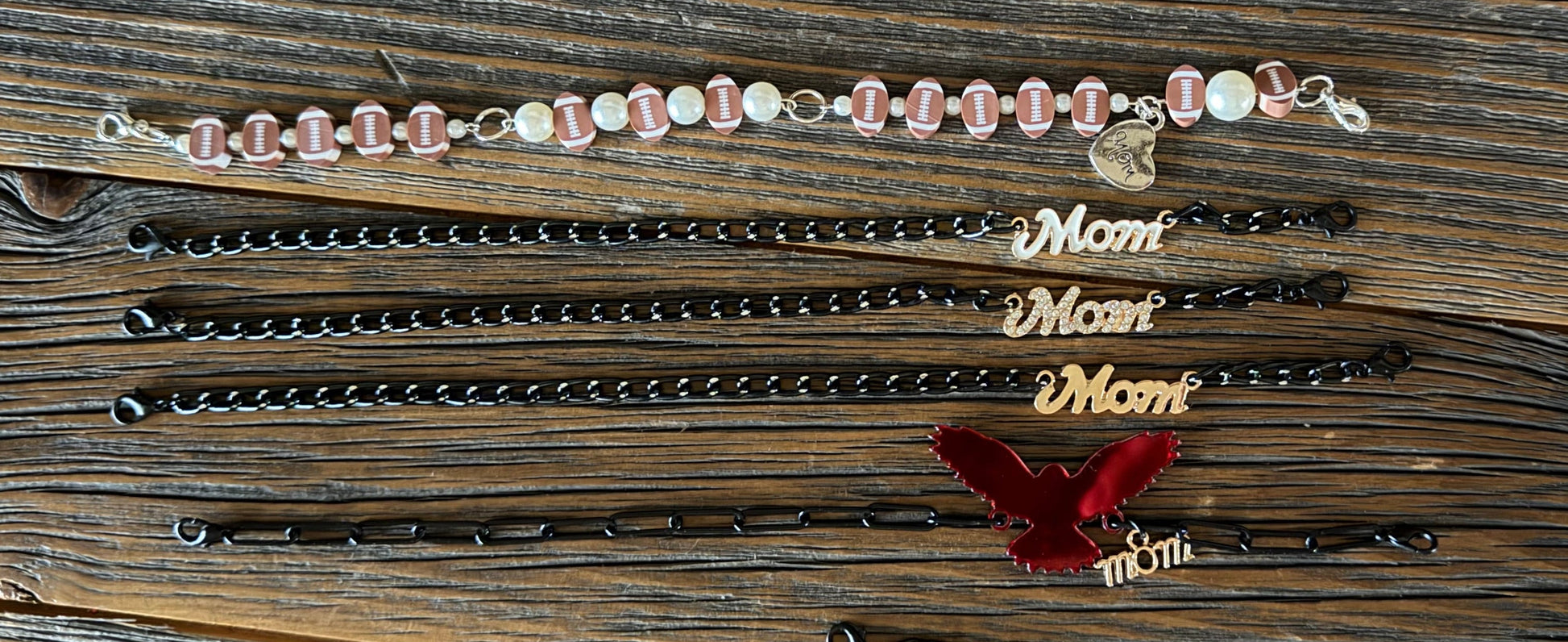 A collection of personalized mother's day bracelets laid out on a rustic wooden plank background. The bracelets include black beaded chains, black links with gold 'Mom' charms, a red velvet ribbon with gold 'Mom' charms, and a pink and white beaded bracelet with a small heart charm.