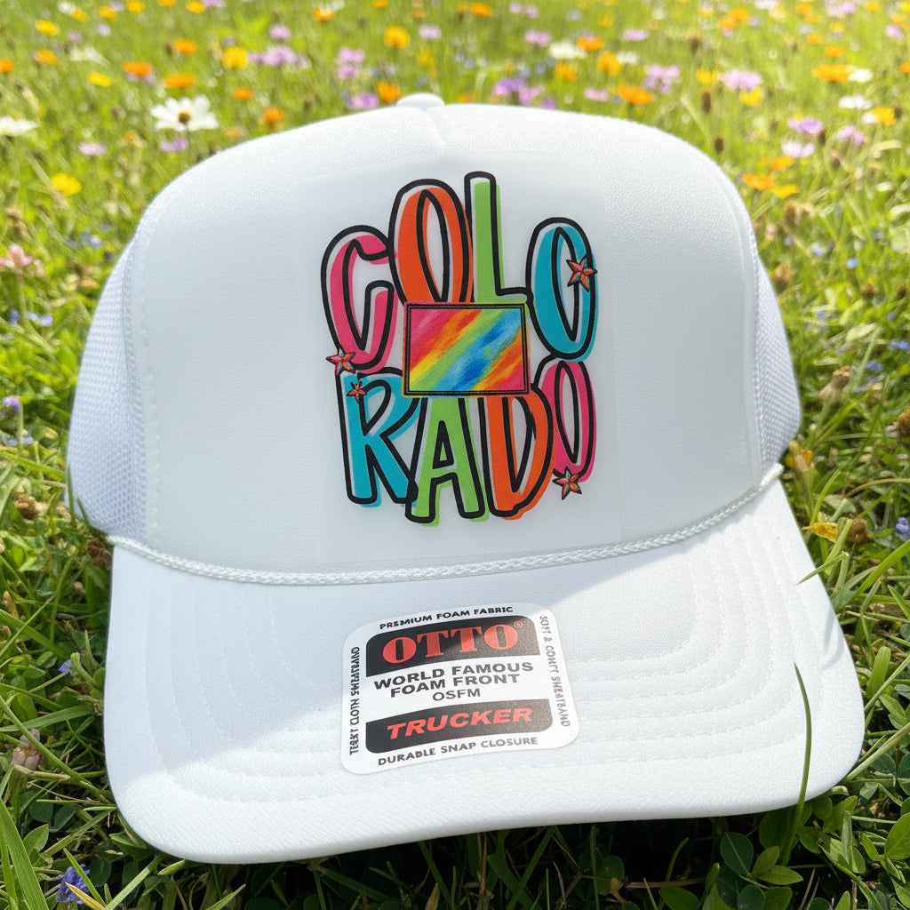 A white trucker hat with a colorful 'COLORADO' graphic on the front, resting on a grassy field with wildflowers. The sticker on the brim shows the brand 'OTTO'.