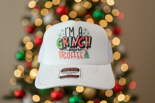 A white trucker hat with a festive graphic that reads 'I'M A GRINCH before COFFEE' in holiday-themed lettering, displayed on a wooden surface.
