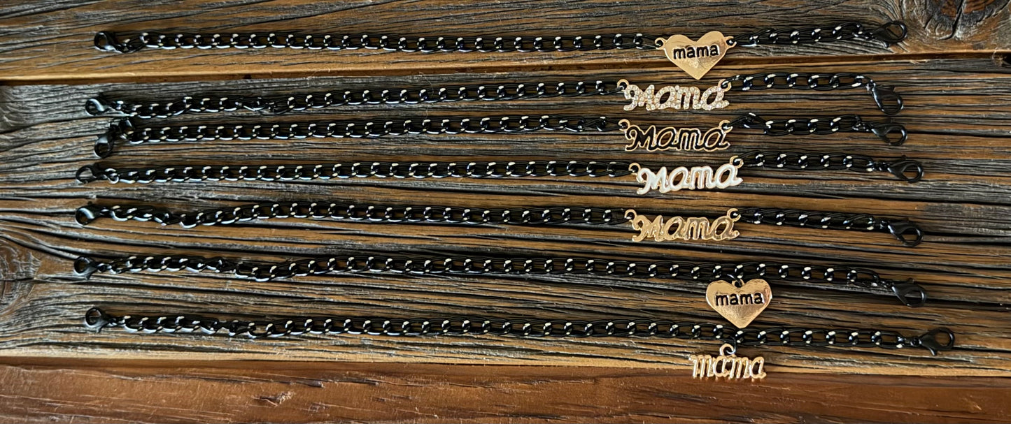 Six black metal link chains with heart-shaped charms engraved with the word 'mama' in gold-tone lettering, arranged on a rustic dark wood background.