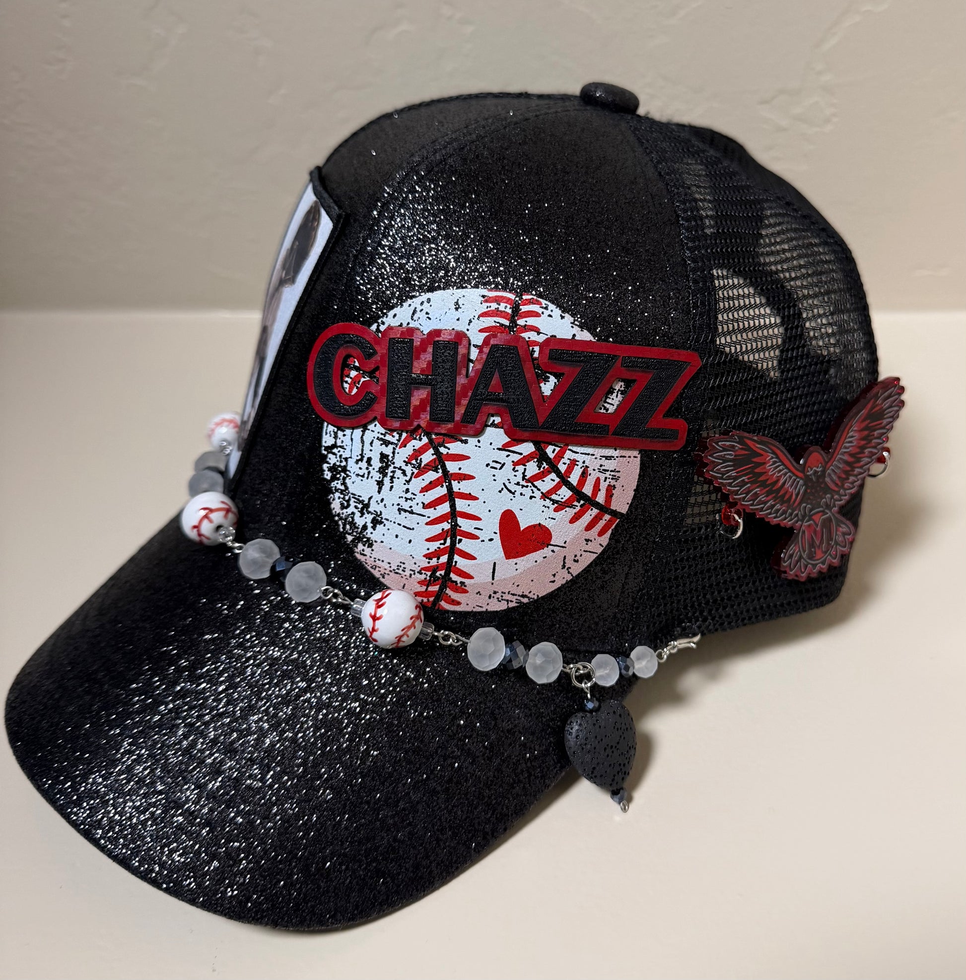 A black glittery trucker cap with a baseball patch, a red 'CHAZZ' text patch, gray beads, and a small bird charm, displayed on a beige surface.