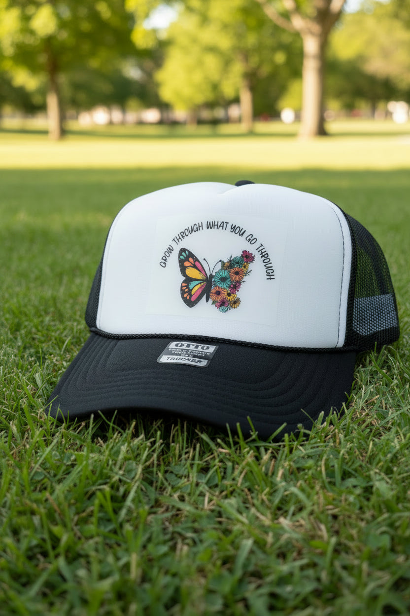A black and white trucker hat with a white front panel, black mesh back, and black brim. The front panel features a colorful butterfly and floral graphic with the text 'DRAW THROUGH WHAT YOU GO THROUGH'.