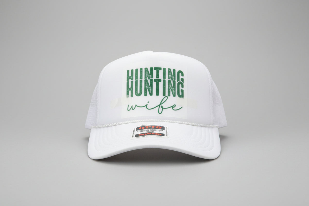 A white trucker hat with a white foam front panel and mesh back, featuring the words 'HUNTING wife' in green on the front. The brim has a visible 'OTC' sticker.