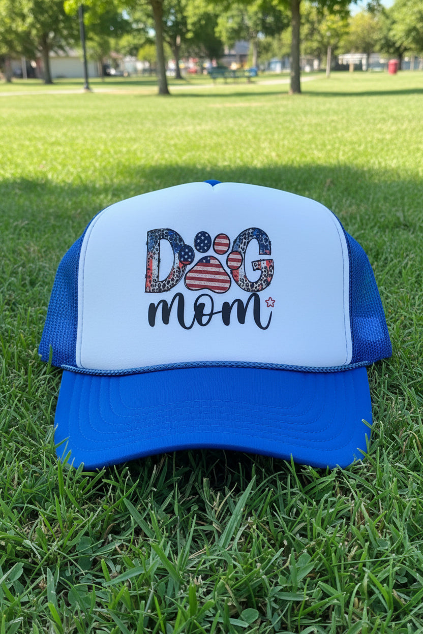 A blue and white trucker hat with 'Dog Mom' written on the front in a patriotic American flag pattern, resting on a wooden table.